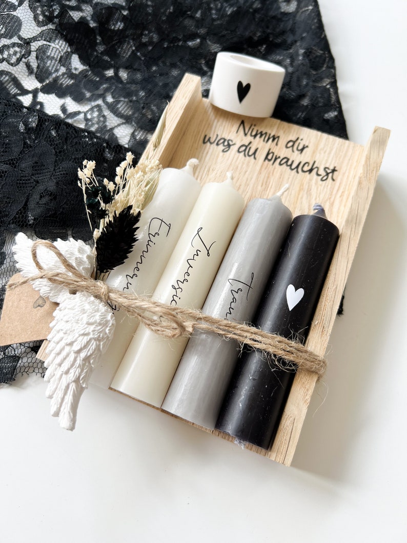 May include: A wooden box with four candles inside. The candles are white, gray, and black. The box is tied with twine and has a small white heart on the side. The text on the box reads "Nimm dir was du brauchst".