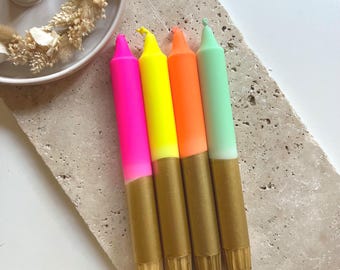 Könnte beinhalten: Vier farbige Stabkerzen mit goldfarbenem Sockel. Die Kerzen sind in Pink, Gelb, Orange und Grün gehalten. Sie sind auf einer strukturierten, hellfarbigen Oberfläche angeordnet. Die Kerzen sind ein dekorativer Artikel.