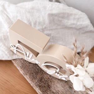 May include: A beige wooden number 50 with the words "Alles Gute" in white script. The number is decorated with dried flowers and sits on a piece of wood. The background is a light-colored fabric.