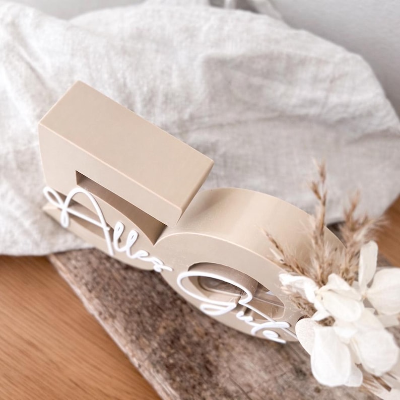 May include: A beige wooden number 50 with the words "Alles Gute" in white script. The number is decorated with dried flowers and sits on a piece of wood. The background is a light-colored fabric.