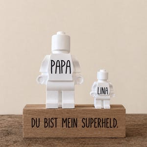 Könnte beinhalten: Zwei weiße Figuren im Lego-Stil, eine große und eine kleine, stehen auf einem Holzblock. Die größere Figur trägt den Aufdruck "PAPA", die kleinere "LINA". Der Block ist mit dem Satz "DU BIST MEIN SUPERHELD" graviert.