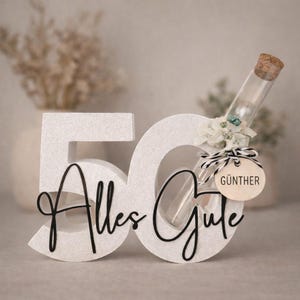 May include: A white glittery "50" with the words "Alles Gute" in black script. A glass vial with a cork stopper is adorned with flowers and a tag that reads "G&uuml;nther".