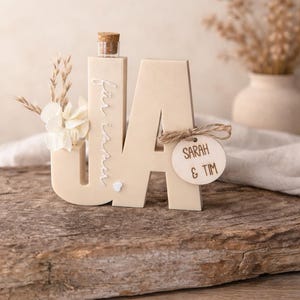 Puede incluir: Un letrero de boda de madera beige con las letras &laquo;JA&raquo;, una peque&ntilde;a botella y decoraciones florales. Se adjunta una etiqueta de madera con &laquo;SARAH & TIM&raquo;. El letrero est&aacute; sobre una superficie de madera, con un fondo neutro.