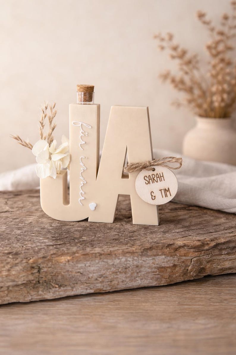 Puede incluir: Un letrero de boda de madera beige con las letras &laquo;JA&raquo;, una peque&ntilde;a botella y decoraciones florales. Se adjunta una etiqueta de madera con &laquo;SARAH & TIM&raquo;. El letrero est&aacute; sobre una superficie de madera, con un fondo neutro.