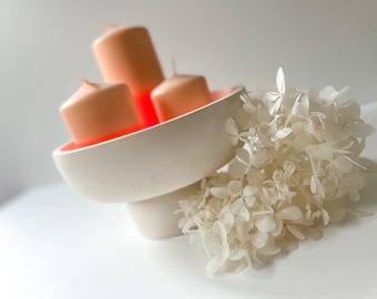 May include: A white ceramic candle holder with three peach-colored candles. The candles are cylindrical and have a small wick. A bunch of white dried flowers is next to the candle holder. The candle holder has a round bowl shape.