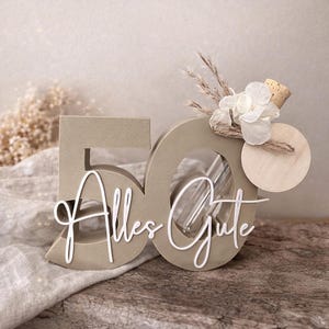 May include: A decorative wooden '50' centerpiece in a neutral beige color, with the cursive text 'Alles Gute' in white. The '0' has a glass tube inside, and is adorned with dried flowers and a wooden disc. A charming birthday decoration.