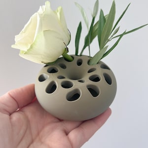 Puede incluir: Un pequeño jarrón en forma de donut, de color topo, con múltiples aberturas circulares. Una rosa blanca y hojas verdes están dispuestas en el jarrón. El jarrón se sostiene en la mano de una persona sobre un fondo blanco.