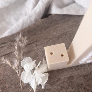 Puede incluir: Un organizador de escritorio minimalista beige con un portal&aacute;pices en forma de cubo con dos agujeros. El organizador est&aacute; acompa&ntilde;ado de flores secas y se encuentra sobre una superficie de madera r&uacute;stica. La est&eacute;tica general es natural y discreta.