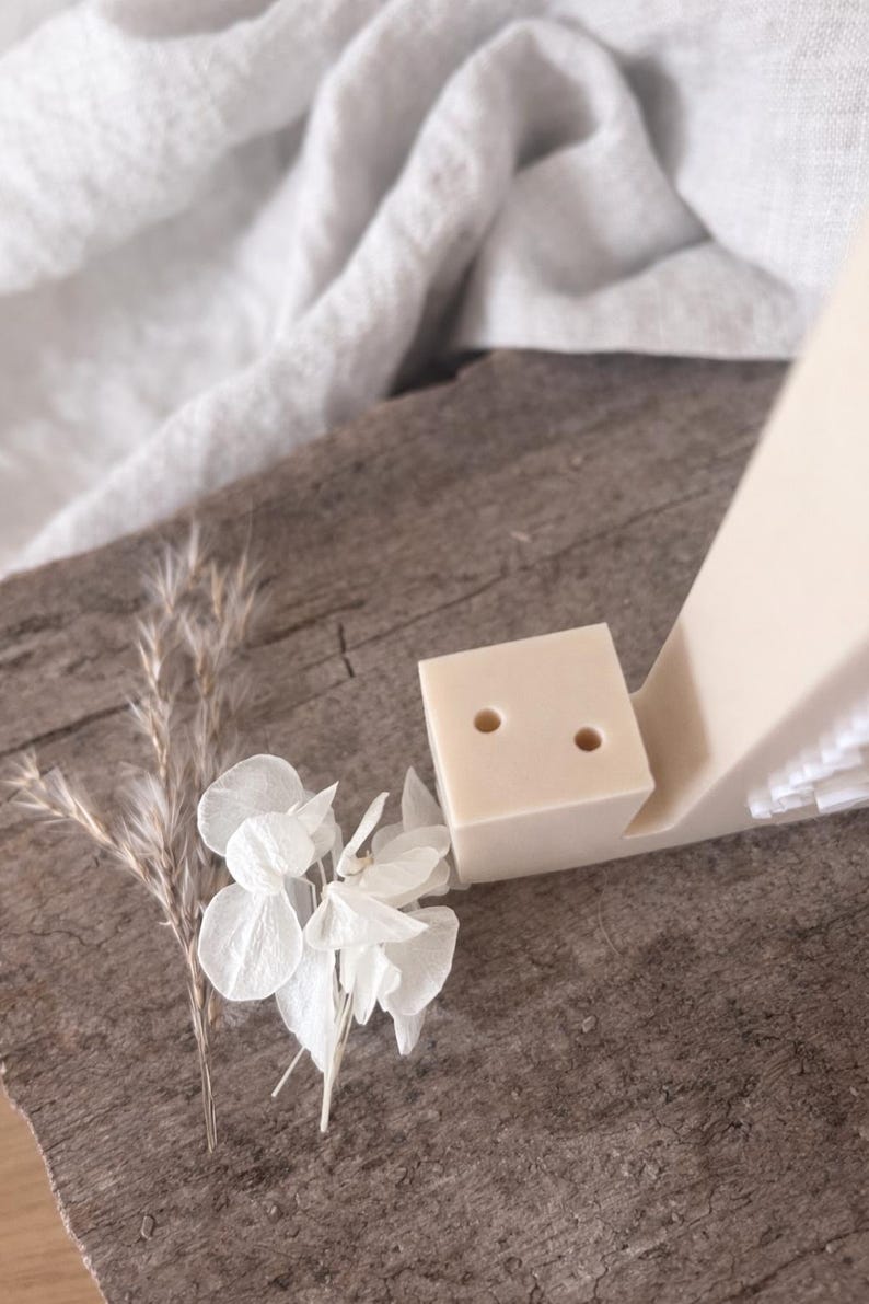 Puede incluir: Un organizador de escritorio minimalista beige con un portal&aacute;pices en forma de cubo con dos agujeros. El organizador est&aacute; acompa&ntilde;ado de flores secas y se encuentra sobre una superficie de madera r&uacute;stica. La est&eacute;tica general es natural y discreta.