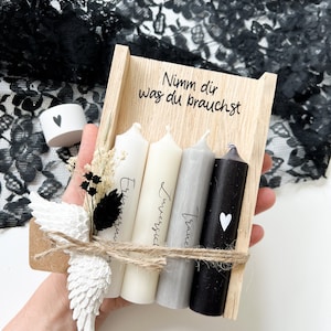 May include: A wooden box with three candles inside. The candles are white, gray, and black. The box is tied with twine and has a small white angel wing attached. The box has the text "Nimm dir was du brauchst" written on it.