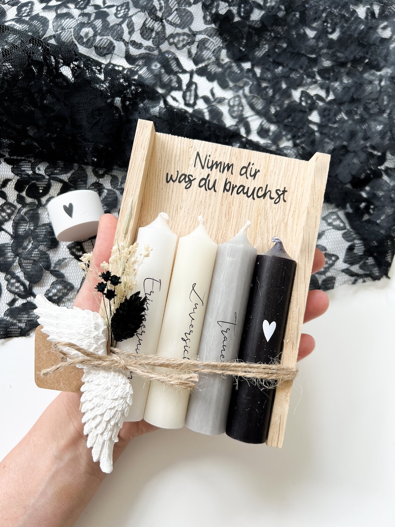 May include: A wooden box with three candles inside. The candles are white, gray, and black. The box is tied with twine and has a small white angel wing attached. The box has the text "Nimm dir was du brauchst" written on it.