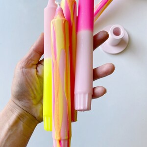 May include: A set of four tall candles with a pink and yellow ombre design. The candles are held in a hand and are ready to be lit.