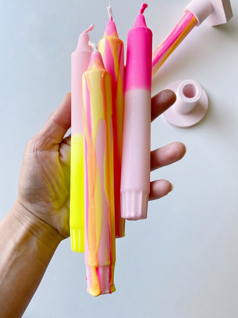 May include: A set of four tall candles with a pink and yellow ombre design. The candles are held in a hand and are ready to be lit.