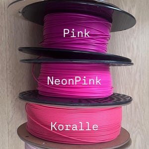 May include: A stack of five spools of 3D printing filament in various shades of pink and red. The colors are labeled "Rot", "Pink", "NeonPink", "Koralle", and "Rosa". The spools are made of cardboard and black plastic.