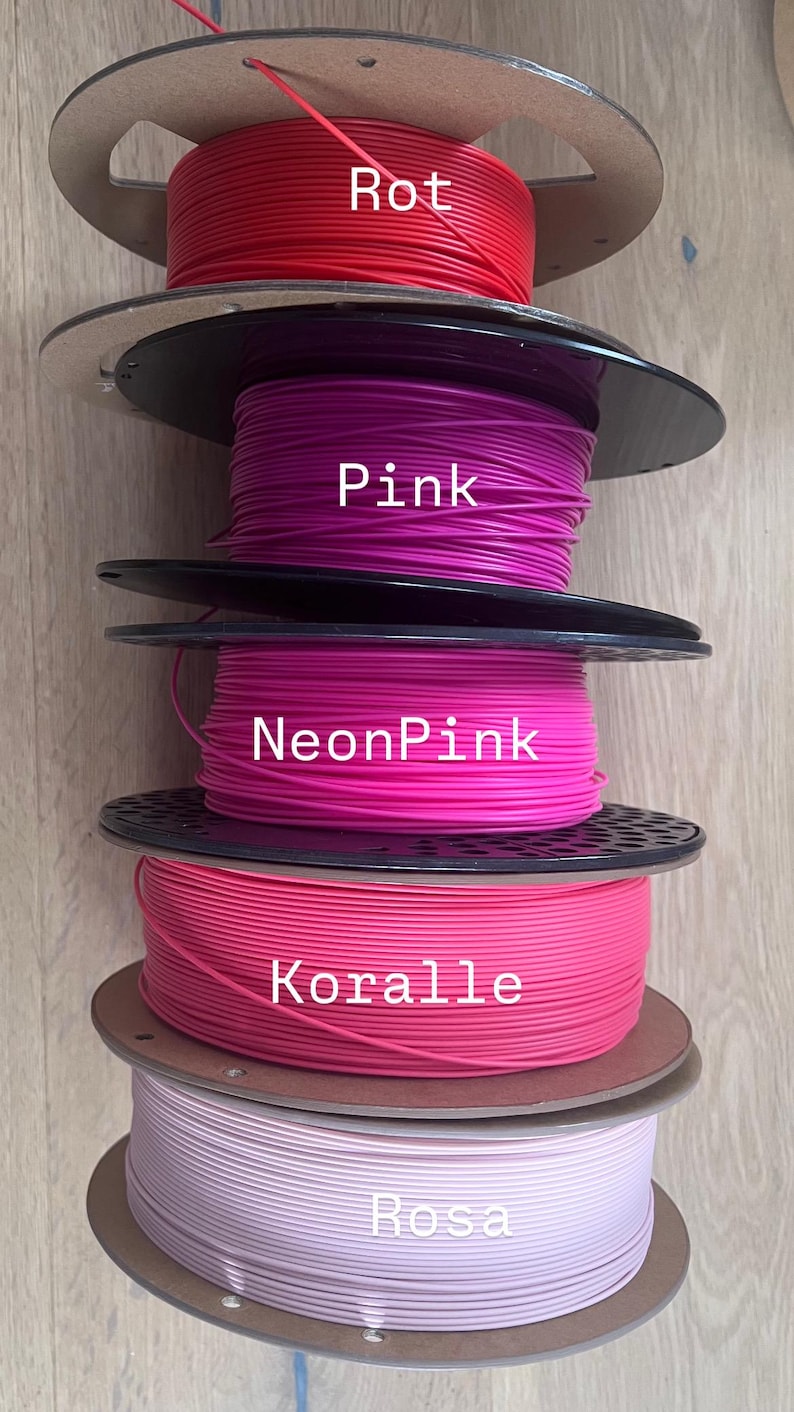 May include: A stack of five spools of 3D printing filament in various shades of pink and red. The colors are labeled "Rot", "Pink", "NeonPink", "Koralle", and "Rosa". The spools are made of cardboard and black plastic.