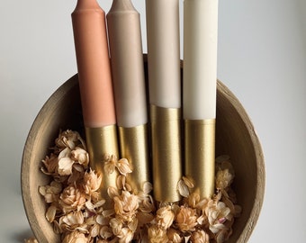 May include: Four candles in a wooden bowl with dried flowers. The candles are in shades of beige, brown, and white with gold accents.