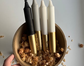 May include: Four candles in a wooden bowl with dried flower petals. The candles are black, white, and gold. The candles are dipped in gold paint.