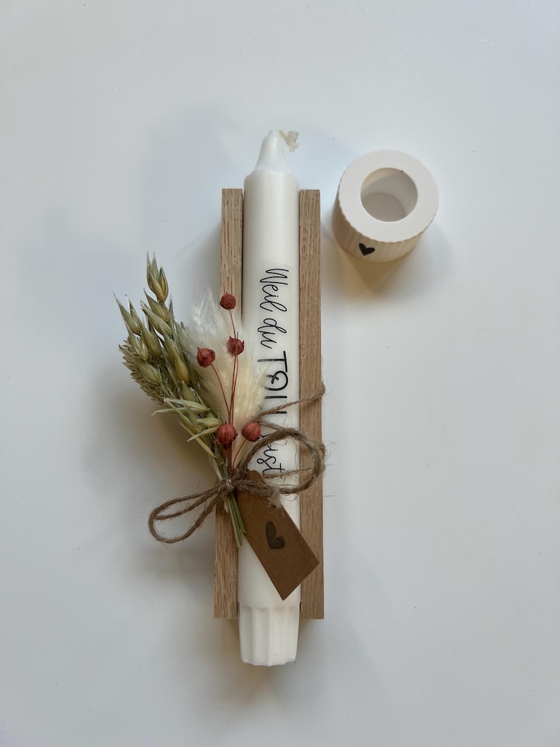 May include: A white candle with the text "Weil du Toll bist" on it, in a wooden holder with a small bouquet of dried flowers and a brown tag with a heart on it.