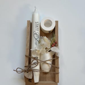 May include: A wooden box with a white candle, a small white vase with dried flowers, and a small white candle holder. The candle has the text "STATT Blau - MP" written on it. The box is tied with twine and has a small wooden heart tag that says "love".