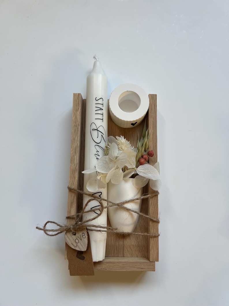 May include: A wooden box with a white candle, a small white vase with dried flowers, and a small white candle holder. The candle has the text "STATT Blau - MP" written on it. The box is tied with twine and has a small wooden heart tag that says "love".