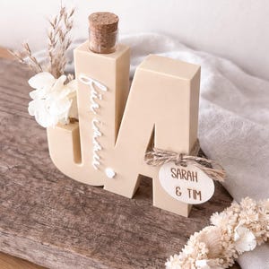 Puede incluir: Una letra "M" de madera beige con una peque&ntilde;a botella y un corcho en la parte superior, y las palabras "f&uuml;r immer" escritas en el lateral. Una peque&ntilde;a etiqueta de madera con "SARAH & TIM" est&aacute; adjunta. La decoraci&oacute;n incluye flores secas.