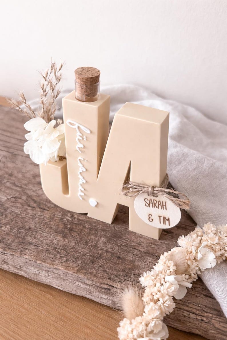 Puede incluir: Una letra "M" de madera beige con una peque&ntilde;a botella y un corcho en la parte superior, y las palabras "f&uuml;r immer" escritas en el lateral. Una peque&ntilde;a etiqueta de madera con "SARAH & TIM" est&aacute; adjunta. La decoraci&oacute;n incluye flores secas.
