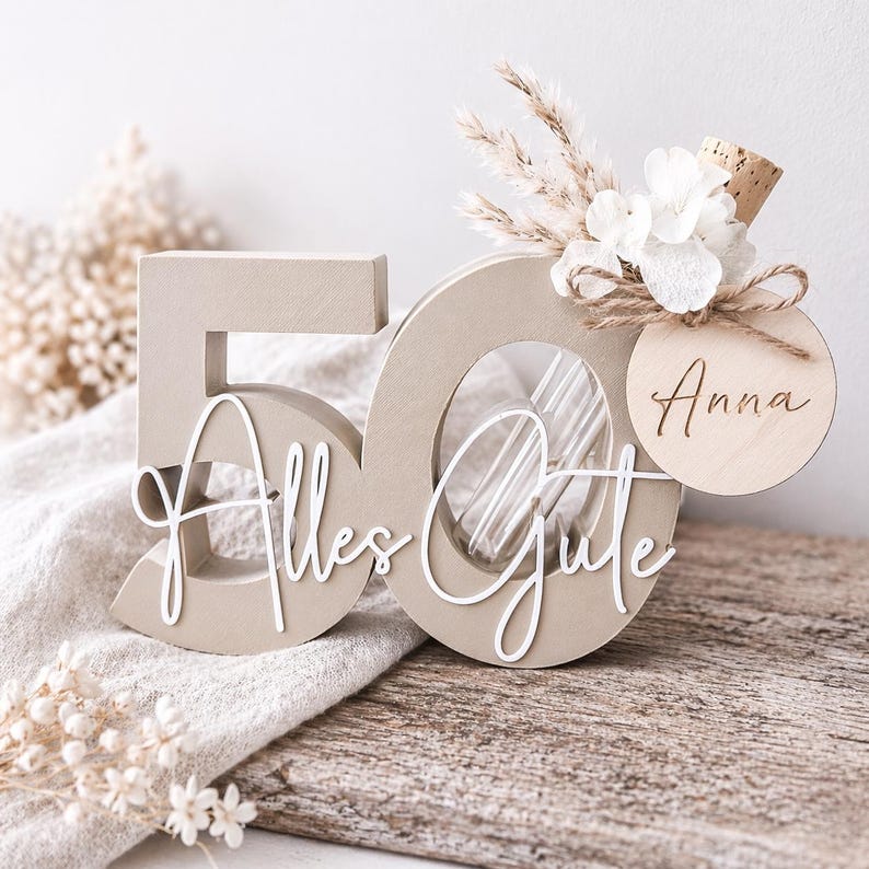 May include: A beige wooden '50' centerpiece with the words 'Alles Gute' in white script. The '0' has a clear insert and is decorated with dried flowers, a cork, and a wooden tag with the name 'Anna'.