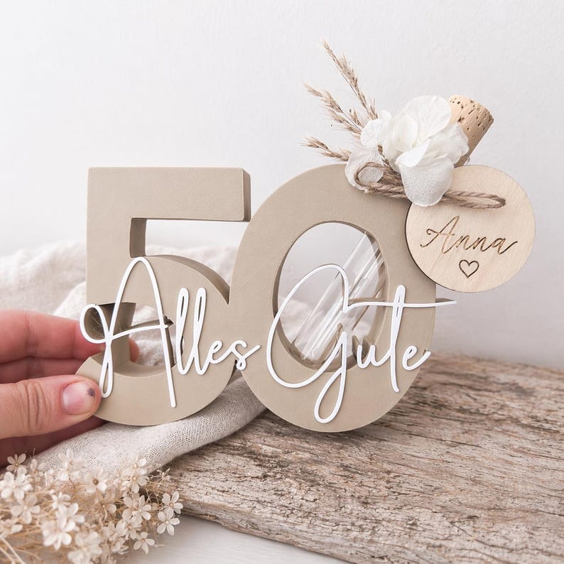 May include: A beige wooden '50' centerpiece with the words 'Alles Gute' in white script. The '0' has a clear insert. Adorned with dried flowers, a cork, and a wooden tag with 'Anna' and a heart. A decorative item.