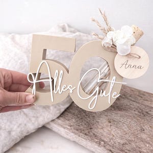 May include: A beige wooden number 50 with the words "Alles Gute" in white script. The number has a clear tube inside and is decorated with flowers, dried plants, and a wooden tag that says "Anna".