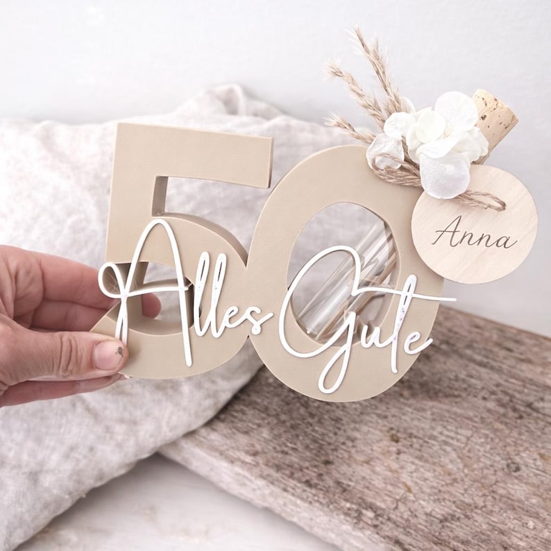 May include: A beige wooden number 50 with the words "Alles Gute" in white script. The number has a clear tube inside and is decorated with flowers, dried plants, and a wooden tag that says "Anna".