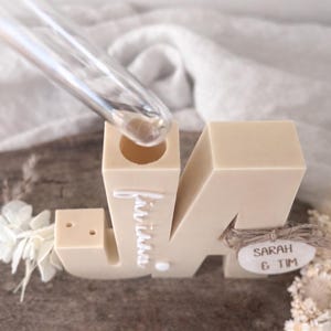 Puede incluir: Un portavelas con forma de letra "M" beige con los nombres "Sarah & Tim" en una etiqueta de madera. La palabra "invitaci&oacute;n" est&aacute; escrita en la letra. Un tubo de vidrio transparente se inserta en la parte superior de la letra. Flores decorativas en el lateral.