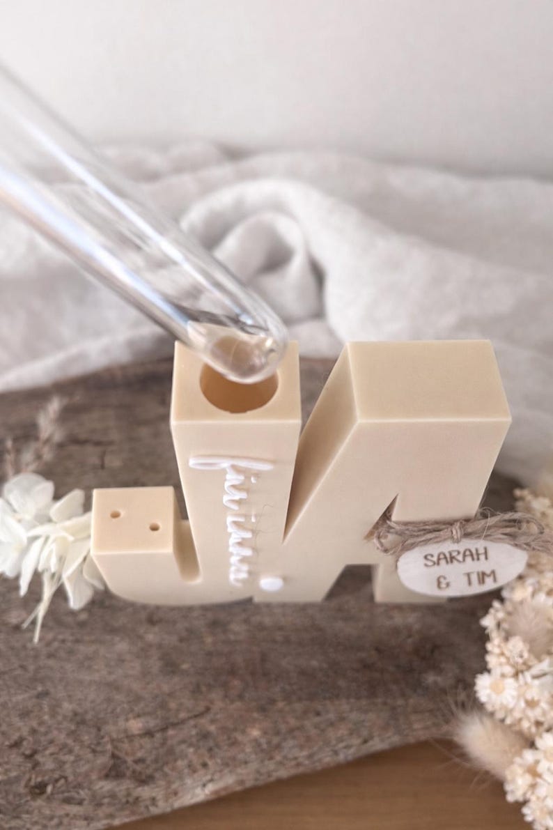 Puede incluir: Un portavelas con forma de letra "M" beige con los nombres "Sarah & Tim" en una etiqueta de madera. La palabra "invitaci&oacute;n" est&aacute; escrita en la letra. Un tubo de vidrio transparente se inserta en la parte superior de la letra. Flores decorativas en el lateral.