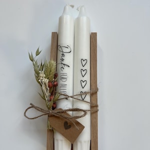 May include: Two white candles with a wooden holder. The candles are decorated with the text "Danke f&uuml;r alles" and hearts. The candles are tied together with twine and a small brown tag with a heart.