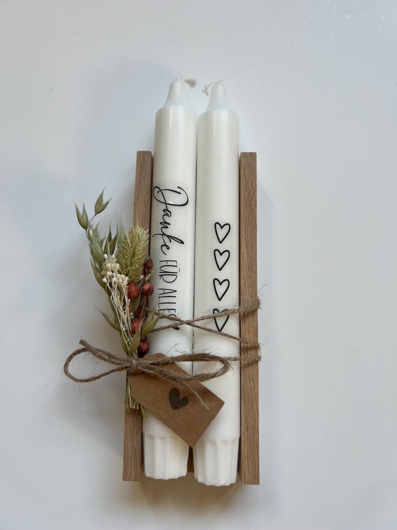 May include: Two white candles with a wooden holder. The candles are decorated with the text "Danke f&uuml;r alles" and hearts. The candles are tied together with twine and a small brown tag with a heart.