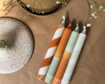 May include: Four taper candles with a striped design in shades of orange, white, and green. The candles are arranged in a row, with the orange and white candles on top and the green and white candles on the bottom.