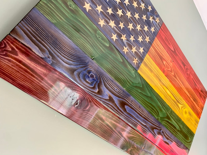 May include: A wooden American flag painted in rainbow colors. The flag is made of reclaimed wood and has a distressed look. The stars are painted gold.