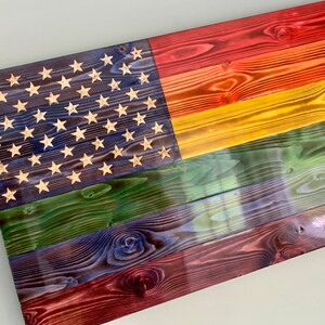 May include: A wooden American flag with a rainbow design. The flag is made of reclaimed wood and has a distressed look. The stars are white and the stripes are red, orange, yellow, green, blue, and purple.