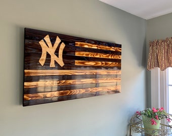 May include: A wooden American flag with the New York Yankees logo in the upper left corner. The flag is made of reclaimed wood and has a distressed look. The flag is hanging on a wall.