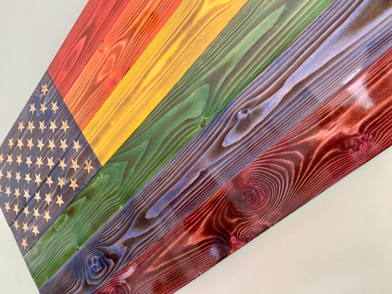 May include: A wooden American flag painted in the colors of the rainbow. The flag is made of individual wood planks stained in red, orange, yellow, green, blue, and purple.