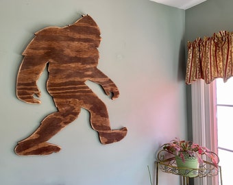 May include: A wooden silhouette of Bigfoot, a mythical creature, walking to the right. The silhouette is brown and has a wood grain texture. It is mounted on a light green wall.