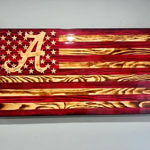 May include: Wooden American flag with a red and natural wood grain finish. The flag features the University of Alabama's "A" logo in the upper left corner, replacing the traditional stars. The stripes are red and natural wood.