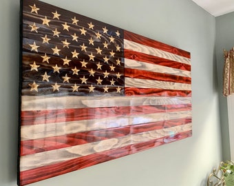 May include: A wooden American flag with red, white, and blue stripes and gold stars on a dark blue background. The flag is hanging on a wall.