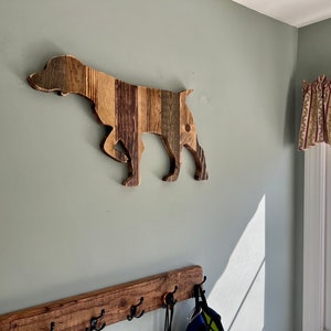May include: A wooden dog silhouette wall decor made from reclaimed wood planks. The dog is in a running pose with its head turned to the side. The wood has a natural brown and gray color with a rustic finish.