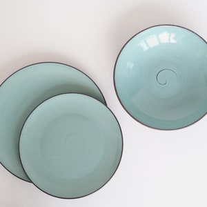 Puede incluir: Tres platos de cerámica de diferentes tamaños, todos con un esmalte azul claro y un borde negro fino. El plato más grande tiene un diseño de espiral sutil en el centro.