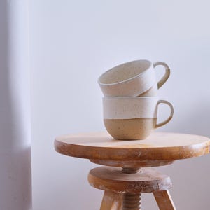 Może przedstawiać: Dwa białe ceramiczne kubki z brązową, kropkowaną glazurą ułożone na drewnianym stołku. Kubki mają zaokrąglony kształt i brązową krawędź.