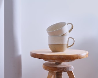 May include: Two white ceramic mugs with brown speckled glaze stacked on a wooden stool. The mugs have a rounded shape and a brown rim.