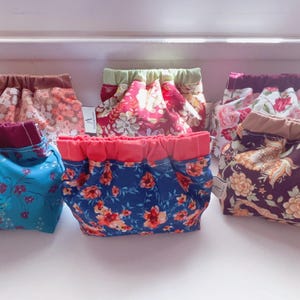 May include: Six small fabric pouches with gathered tops and floral patterns in various colors including blue, red, pink, and brown. Each pouch features a solid-colored fabric band at the top opening and a small white tag attached to the side.