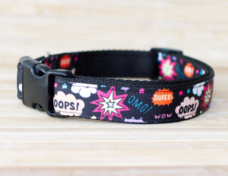 May include: Black dog collar with a comic book-style pattern. The collar features a black plastic buckle and a repeating design of speech bubbles with words like "OOPS!" and "OMG!", along with starburst graphics in pink, orange, and white.
