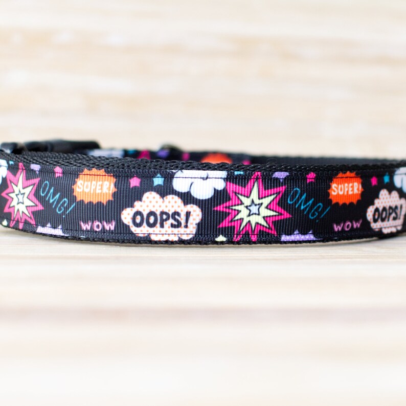 May include: Black dog collar with a comic book style design. The collar features colorful speech bubbles with words like "OMG!", "SUPER!", and "OOPS!" along with starburst and star graphics. The collar has a black plastic buckle.
