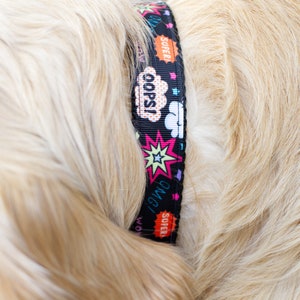 May include: Black dog collar with comic book style graphics. The collar features the words "Oops!", "OMG!", and "Super!" in speech bubbles and starbursts. The collar is on a dog with light brown fur.